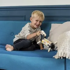 A smiling young boy sits on a blue couch, playing with a wooden toy dog with a segmented body.