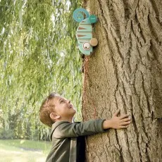 A happy boy hugging a large tree with a Janod chameleon toy attached to the trunk.