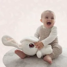 A happy baby smiles broadly while playing with a soft white rabbit toy on a gray circle rug.