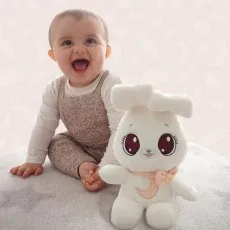 A joyful baby sits next to a white plush bunny with big pink eyes, both looking towards the camera.