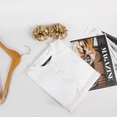 Folded white eyelet boho blouse on a white surface with gold scrunchies, a wooden hanger, and magazines..